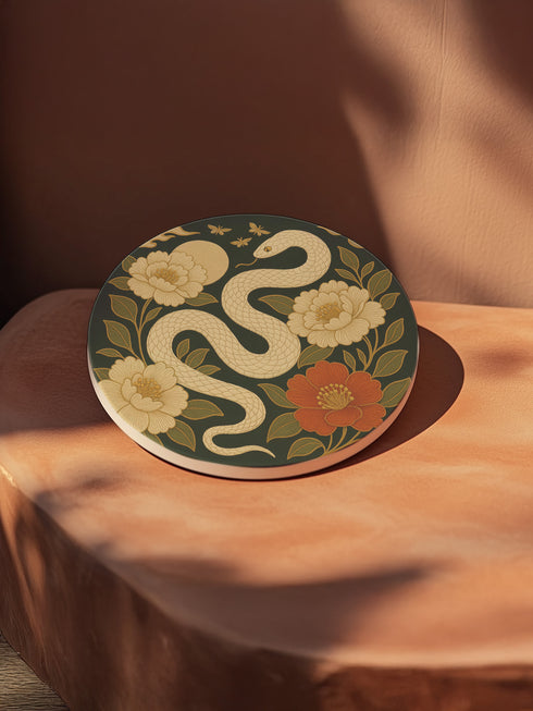 Round ceramic coaster with a chinoiserie-inspired design featuring a white snake among golden peonies, butterflies, and a glowing moon on a dark green background. Surrounded by floral and botanical accents with a soft vintage aesthetic. The coaster has a cork back and measures about 4 inches in diameter. Ideal for mugs, teacups, or wine glasses, this decorative piece suits witchy, victorian, and chinoiserie-inspired decor.